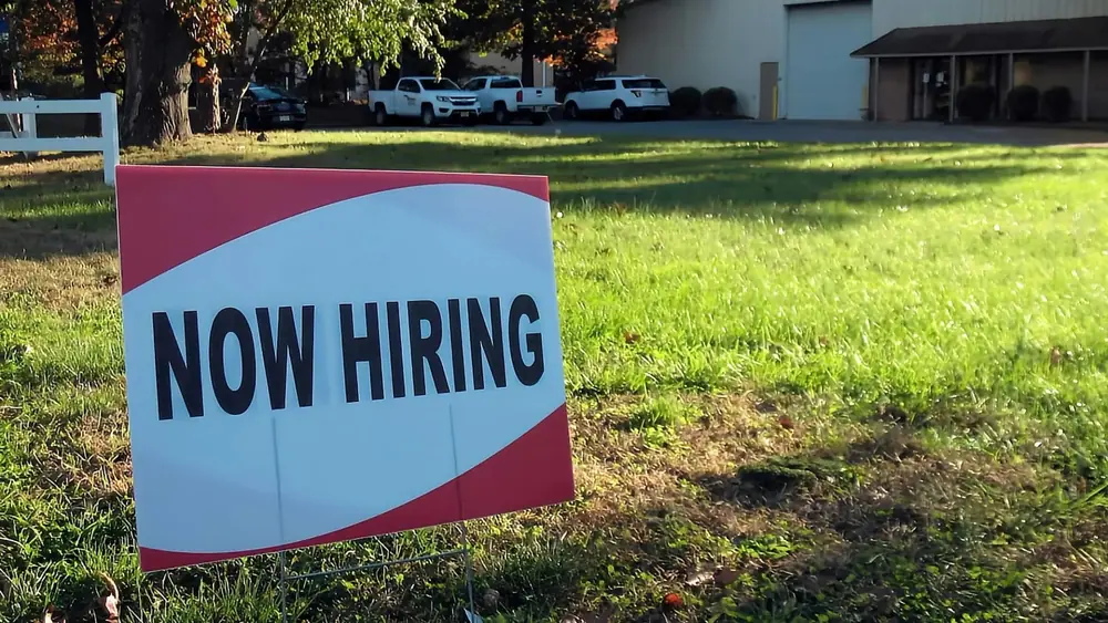 Sign saying "Now Hiring" sits on a lawn.