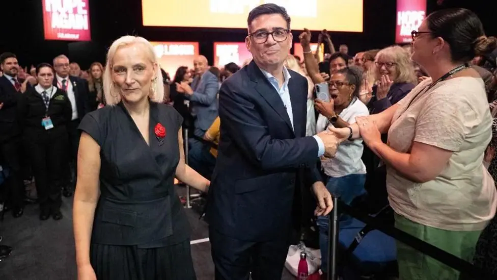 Marie-France van Heel and Andy Burnham greet delegates in the audience after the prime minister's keynote speech. Van Heel is wearing a dark-coloured dress with no sleeves. Burnham is wearing a…