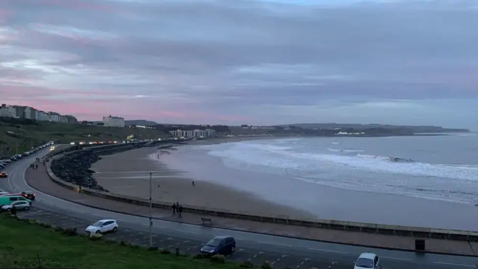 This is an early evening shot of Scarborough's North Bay. The sky is a striking mix of purple and pink. The tide is out and a small number of people are walking on the beach. Traffic is light along…