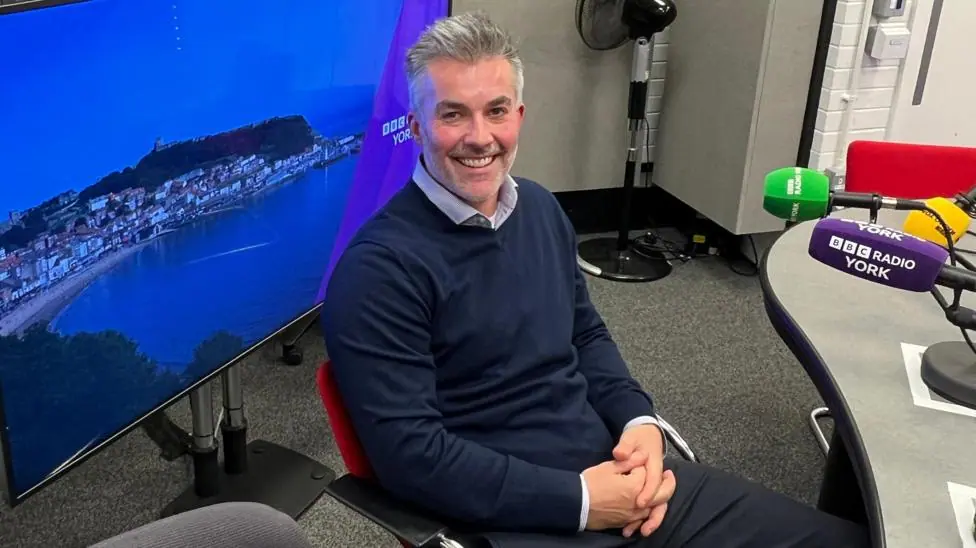 Labour mayor of York and North Yorkshire, David Skaith, is sitting in the BBC Radio York hotseat, waiting to be interviewed by presenter Joanita Musisi. He is smiling at the camera, wearing a blue…