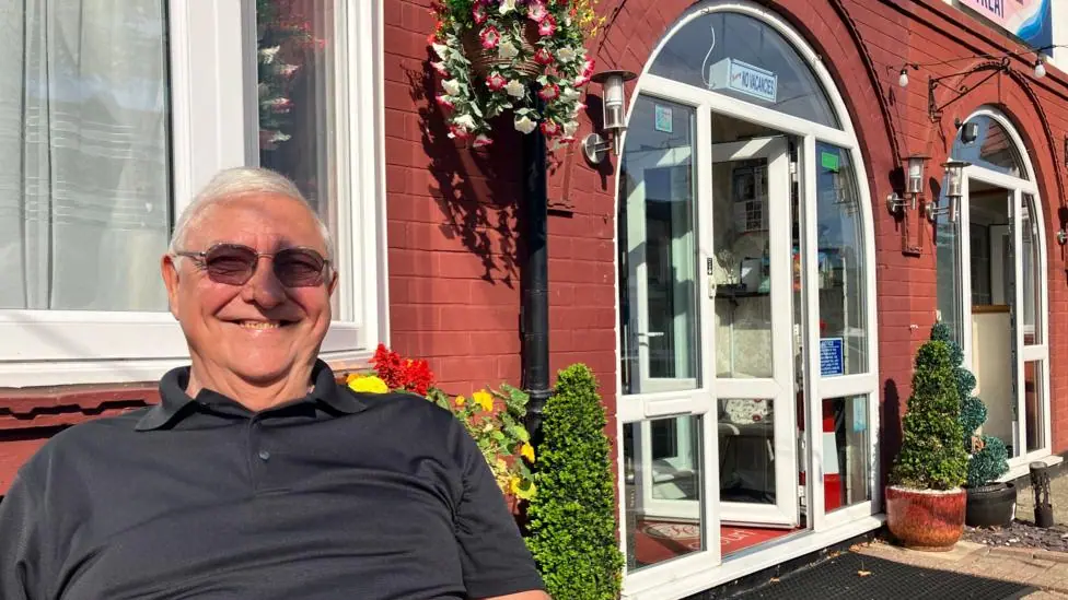 A man sitting in the sunshine outside a B&B in Scarborough. He is smiling at the camera. The sun is on his face and a hanging basket, full of pink and white flowers, is attached to a lamppost behind…