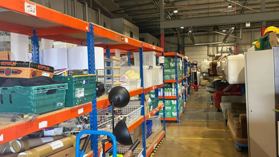 Inside the warehouse there are shelving units with boxes on them, in the background some have long life milks on the shelves and sofas are piled in another row.