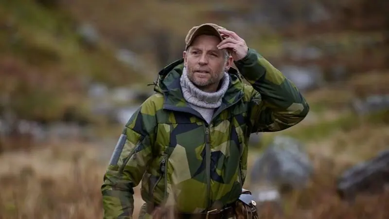 Andrew Price is wearing a green camouflage jacket with a grey turtleneck surrounded by plants and grass.