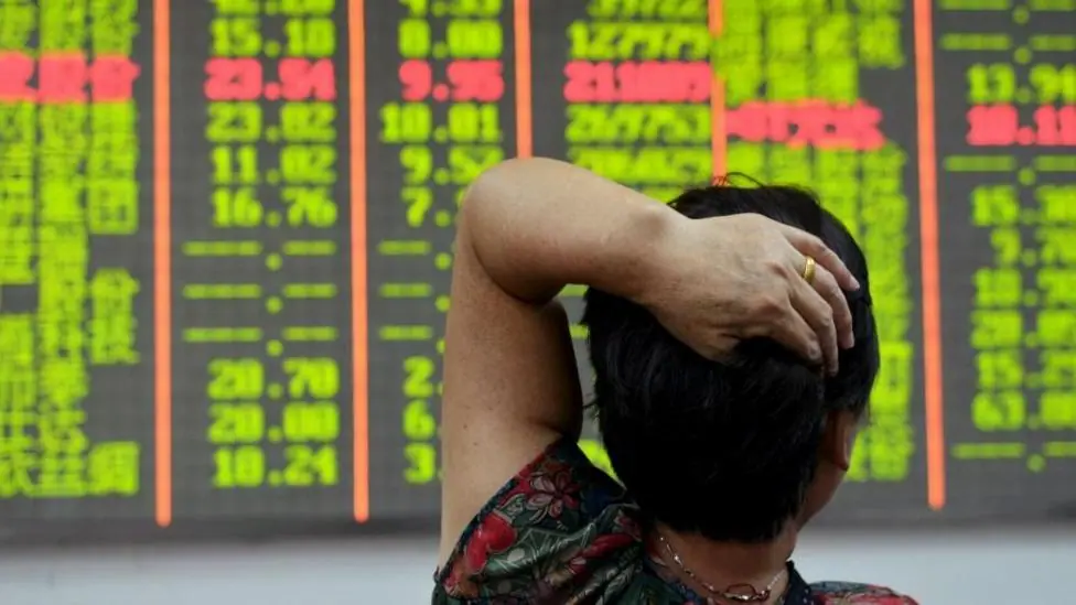 An investor looks at an electronic board showing stock information with numbers highlighted in red and lime green against a black background. The investor has her back to the camera and has her left…