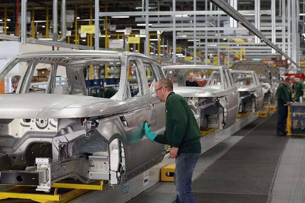 A factory line of shells of cars- a man wearing a green jumper, safety goggles and a green glove is standing sideways working on one of the cars. Colleagues can be seen in the background also wearing…