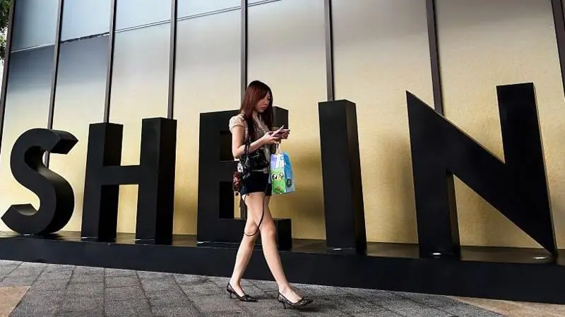 A woman looks at her smartphone while walking past a SHEIN sign outside the company's office on 14 July, 2026, in Guangzhou, Guangdong Province, China.
