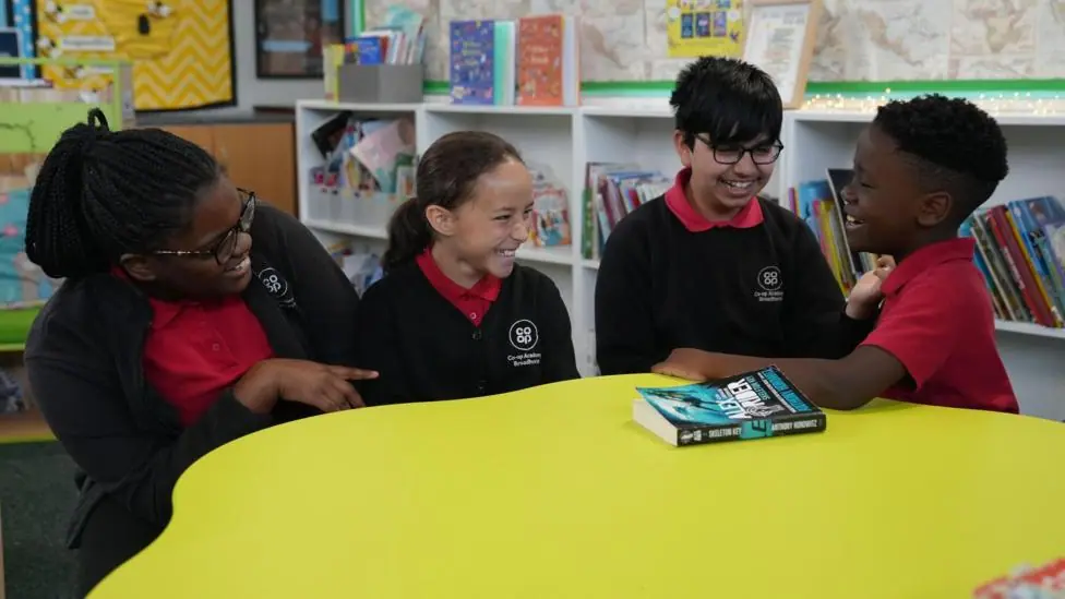 Four primary-age pupils sit around a large circular yellow table in a school library or reading area. The pupils are wearing red polo shirts with dark cardigans or jumpers.