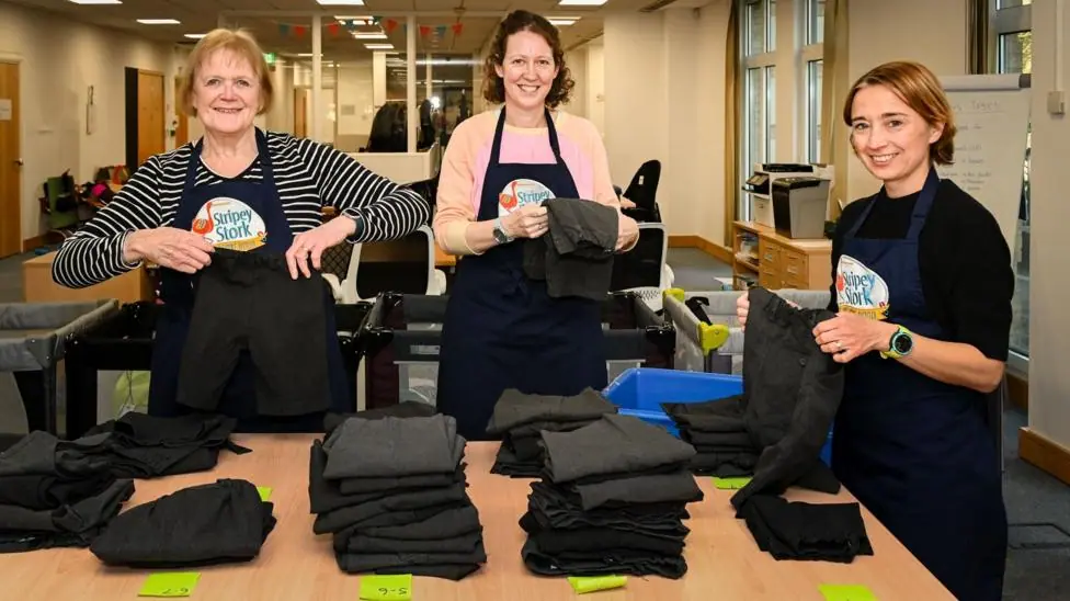 Three workers at Stripey Stork folding uniform items to be administered to children in Surrey. The three women are all wearing navy aprons with the charity's logo, and are all stood beside piles of…