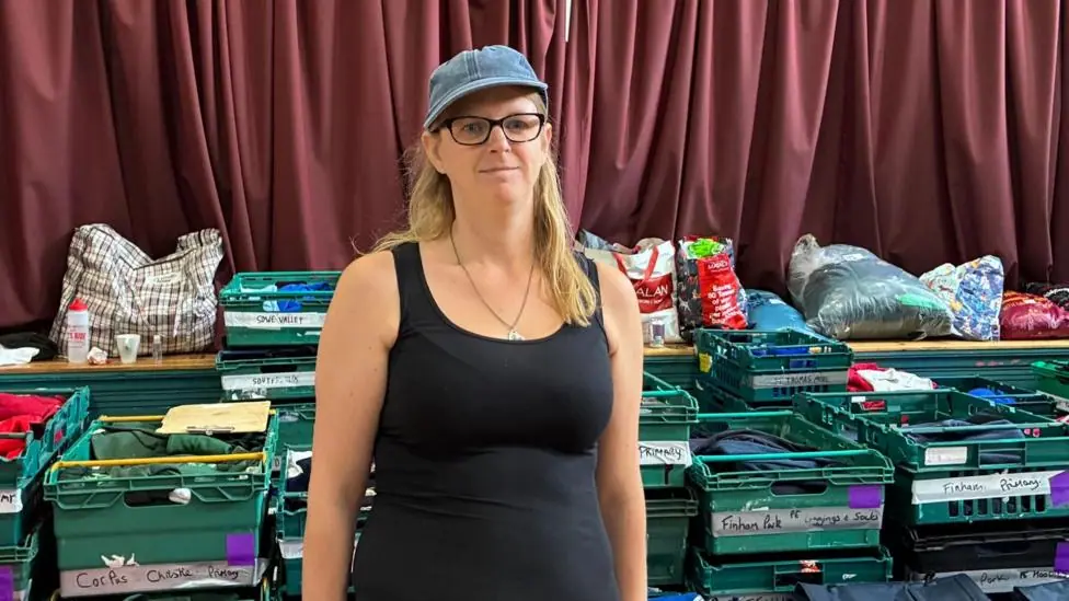 Zia Lee stands in a black vest top with a denim cap on and black glasses and long blonde hair. Behind her are green crates with school names and carefully folded uniforms in them.
