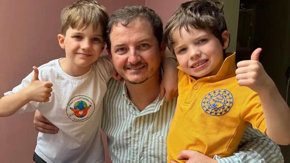 Jon who is 42 is pictured with his two children 10 year old Tom and six year old, Sion in amongst several blue school jumpers and several yellow polo shirts from Ysgol Treganna primary school