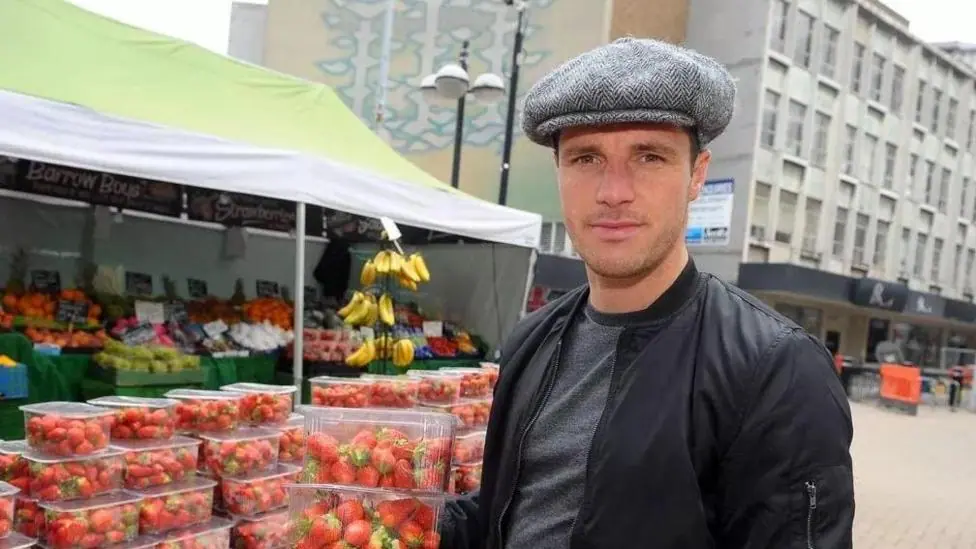 Tommy Coyle wearing a flat cap, standing next to the fruit stall in Hull City Centre. He is holding multiple packs of strawberries and is wearing a black zip up jacket.