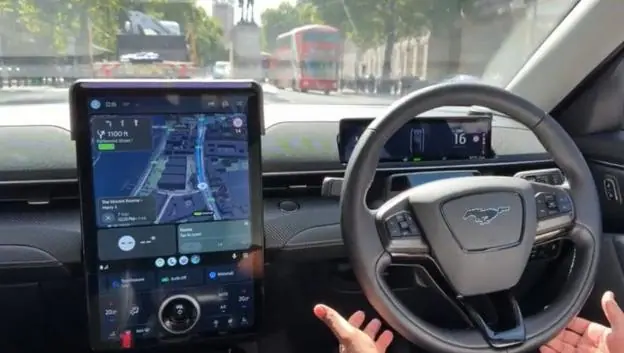 A passenger's view inside a robotaxi as it drives through London, with the hands of the safety driver not touching but around the wheel of the car, and a large black tablet with a map on it showing…