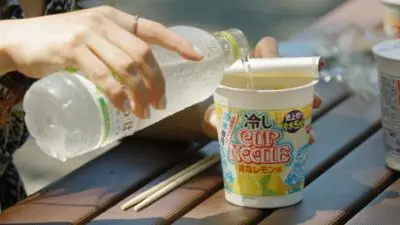 Woman's hand pouring water from a bottle into a cup noddle sitting an a wooden table outside