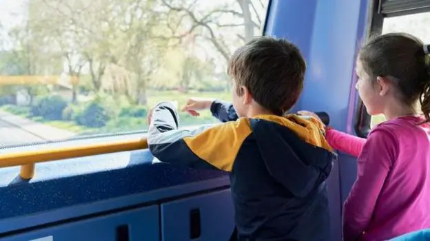 An image of a two children aged around 7 looking out of the front window on the upstairs of a bus. One has a brown ponytail and is wearing a pink long-sleeved top, the other has short brown hair and…