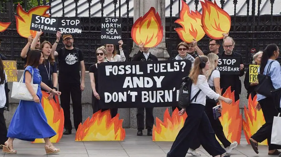 Climate activists stand outside a wrought-iron fence holding flame-shaped placards and signs opposing North Sea oil and gas projects, including Rosebank and Jackdaw, while pedestrians walk past in…