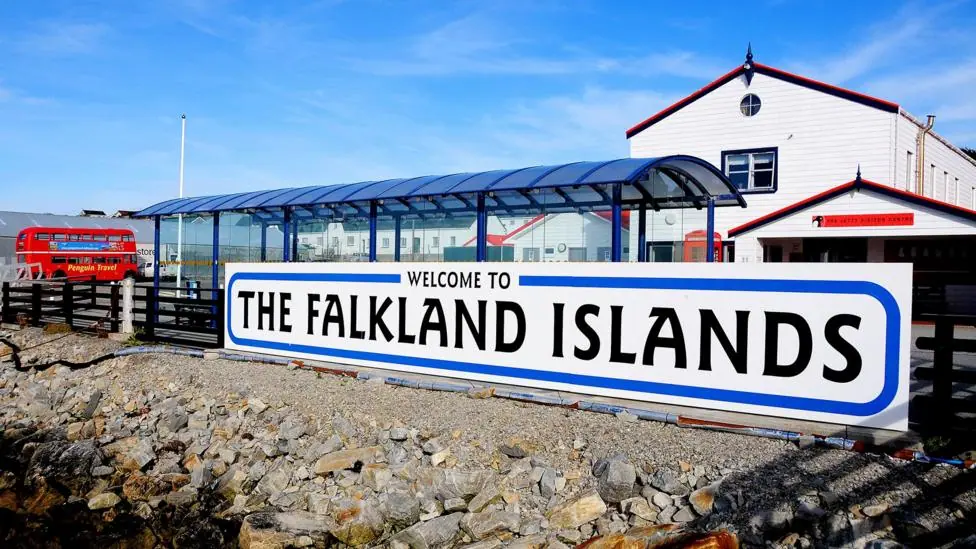 A sign saying 'welcome to The Falkland Islands' in front of a bus terminal at the harbour at Port Stanley in the Falklands Islands, with a red London-style bus in the background against a clear blue…
