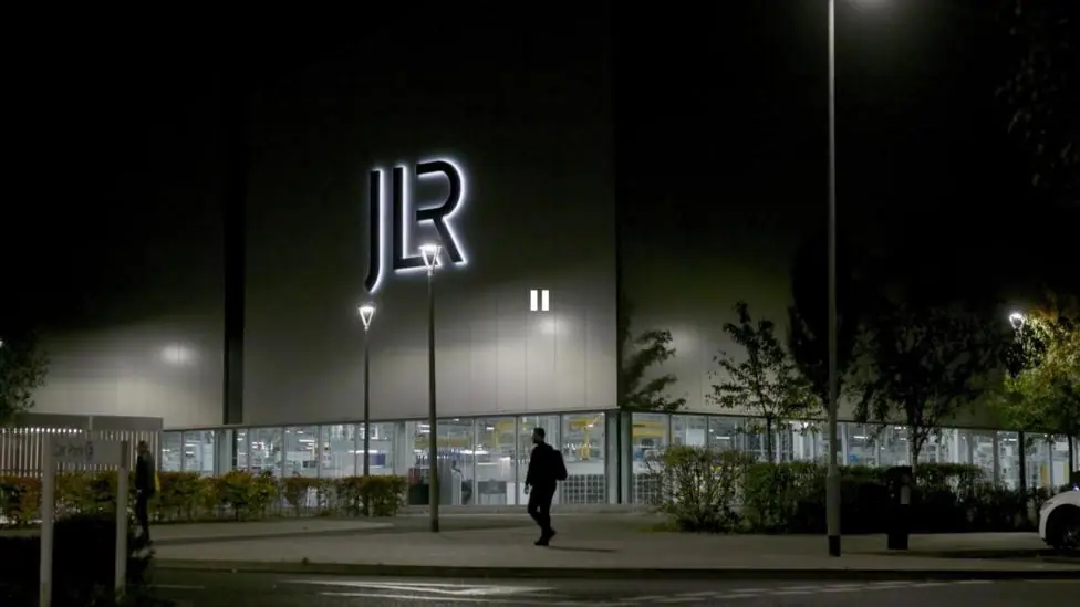 The exterior of Jaguar Land Rover's Wolverhampton factory. It is pictured at night, with a silhouetted person walking in front of the illuminated frontage of the building.