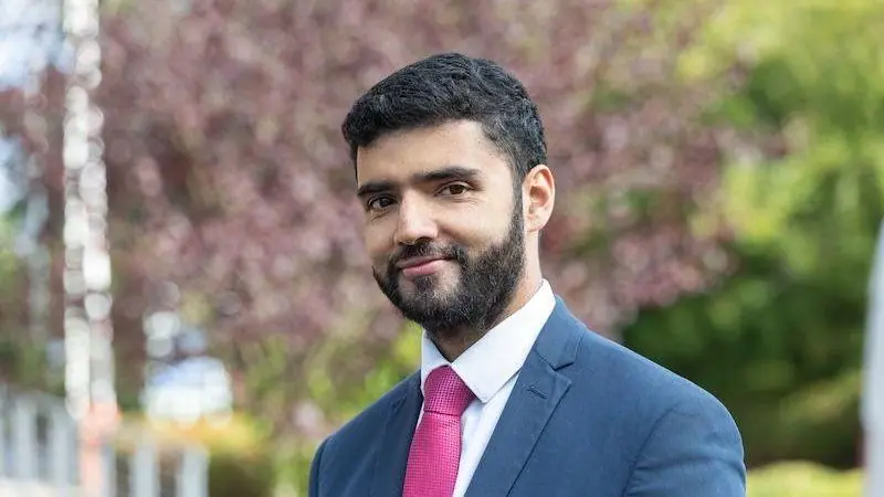 A man with short black hair and a beard is smiling at the camera. He is wearing a blue suit, a white shirt and a pink tie and is standing outdoors with greenery in the background that has been…