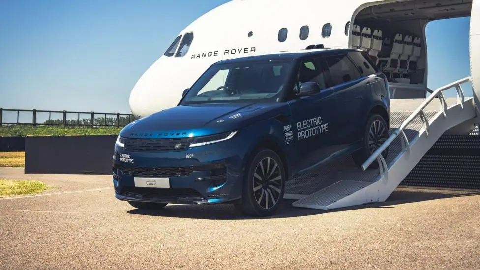 A blue Range Rover Electric prototype drives down a metal ramp from the open cargo bay of a large white aircraft. The vehicle carries “Electric Prototype” markings and technical angle indicators on…