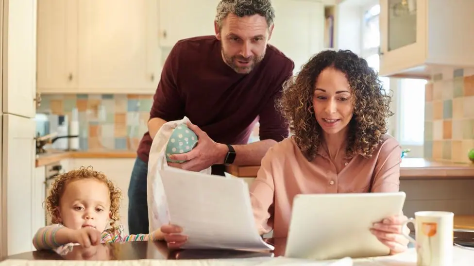 A family of three looking at bills and finances at a kitchen table. From left to right, a child is standing on the floor looking out across the table, a man is looking over a woman's shoulder at A4…