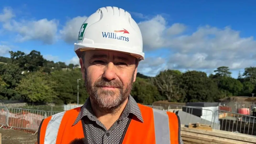 A picture of a man wearing a white hard hat and a bright orange high vis jacket. He's looking at the camera and standing on a building site. There are fences and trees behind him