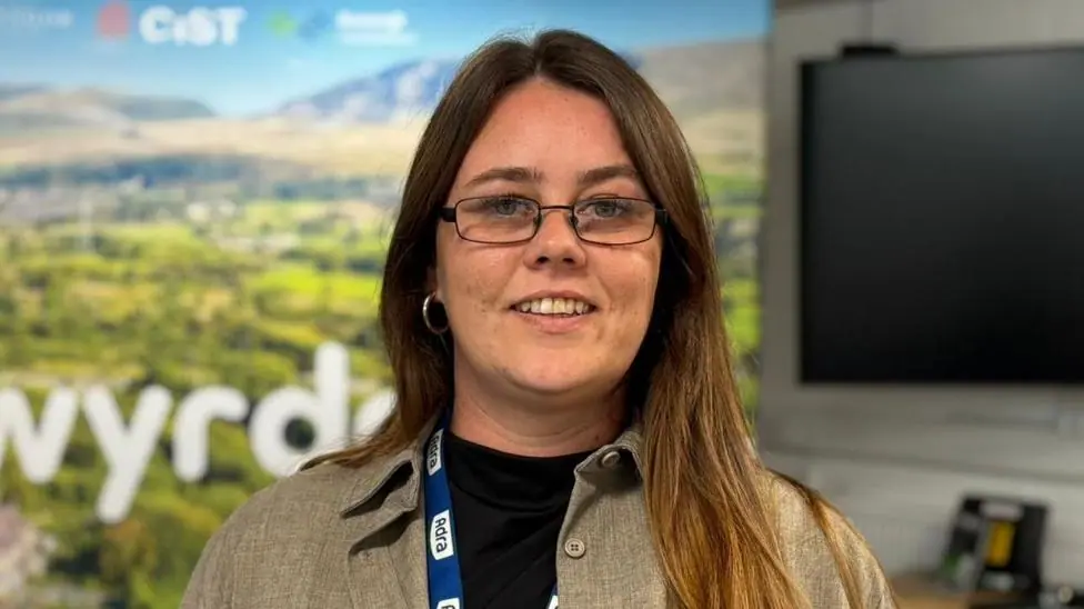 A picture of a woman wearing a light brown coloured shirt and a black tshirt. She's wearing glasses and a lanyard around her neck. Behind her is a TV that is turned off and a large sign showing…