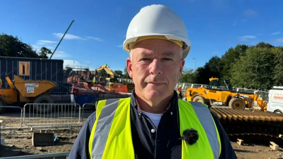 A picture of a man wearing a white hard hat and a bright yellow high vis jacket. He's looking at the camera and standing on a building site. There are fences and heavy machinery behind him including…