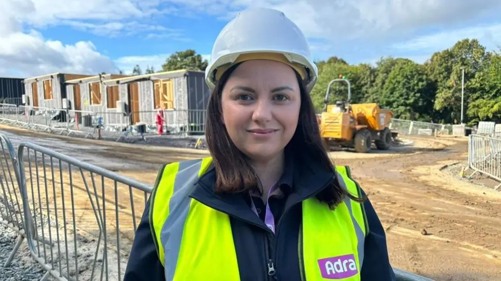 A picture of a woman wearing a white hard hat and a yellow high vis jacket. She's looking at the camera and standing on a building site. There's a dumper truck behind her and also homes that are…
