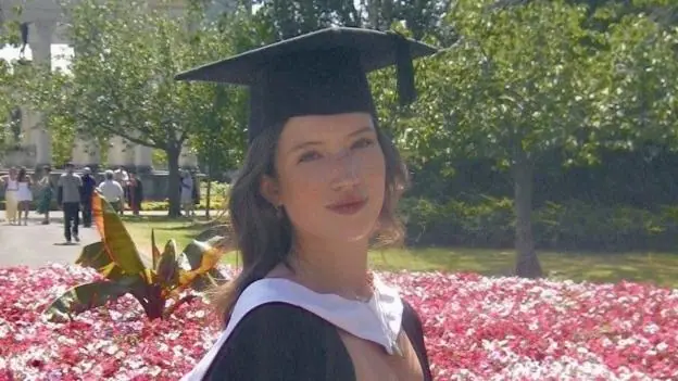 A girl with curly dark hair, wearing a white dress and black graduation cap and gown. She is stood in front of a flowerbed of pink flowers, with trees and people in the background, and is smiling at…
