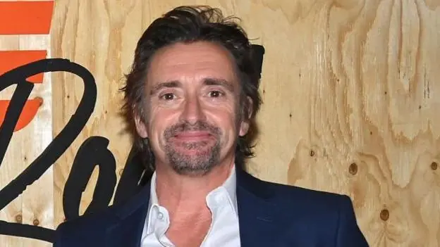 Richard Hammond is photographed from the shoulders up, standing in front of a promotional sign for the TV series 'The Grand Tour'. His face is facing slightly to the side and he is smiling at the…