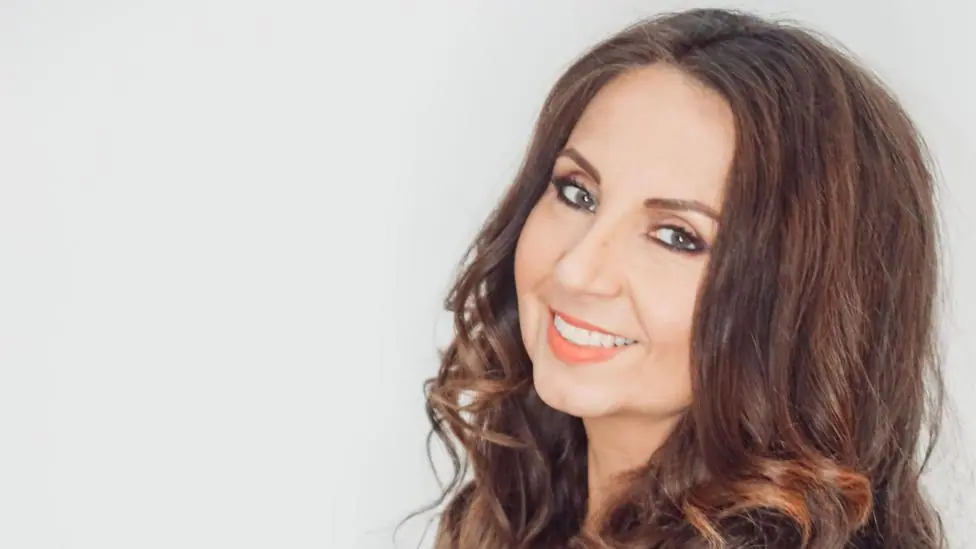 Melissa Alcruz with long brown wavy hair smiles at the camera, against a light background