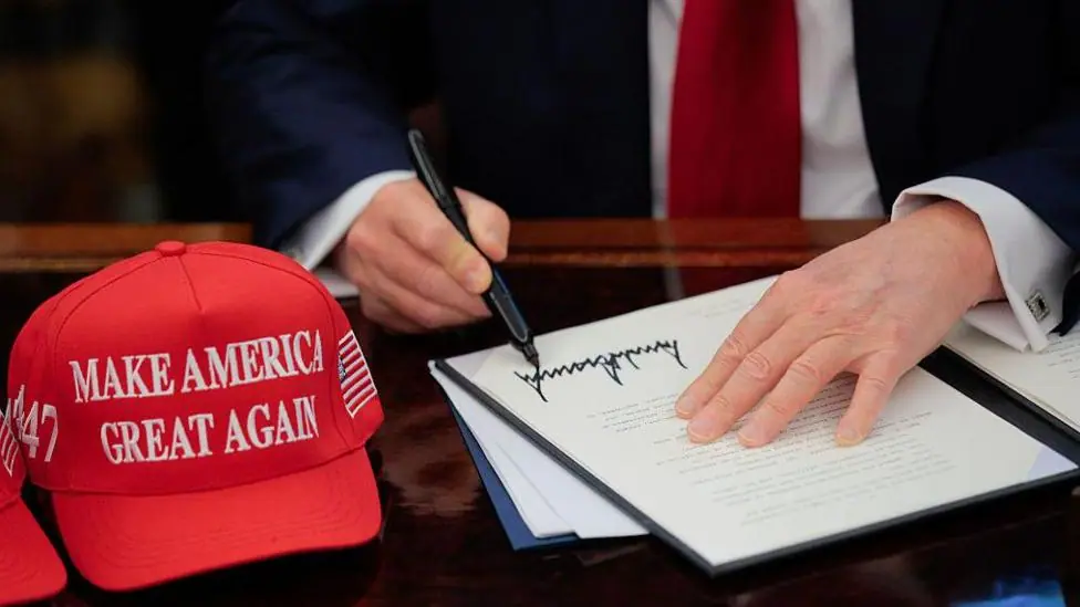 US President Donald Trump signs an executive order in the Oval Office at the White House in Washington, DC on 23 April 2025. A red baseball hat embroidered with "Make America Great Again" in white…