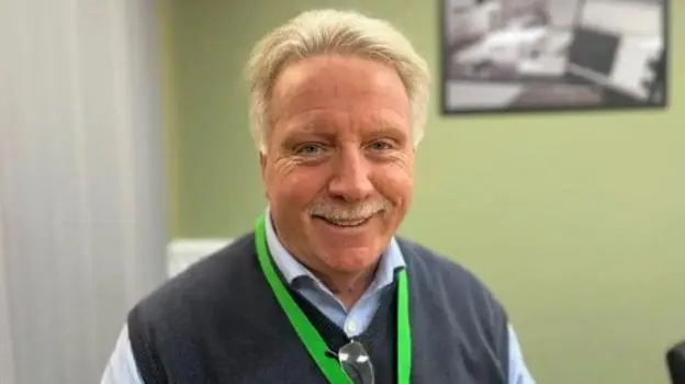 A man with blonde hair and a blonde moustache smiles into the camera. He is wearing a grey pullover with a shirt underneath and a bright green lanyard. There is a green wall behind him