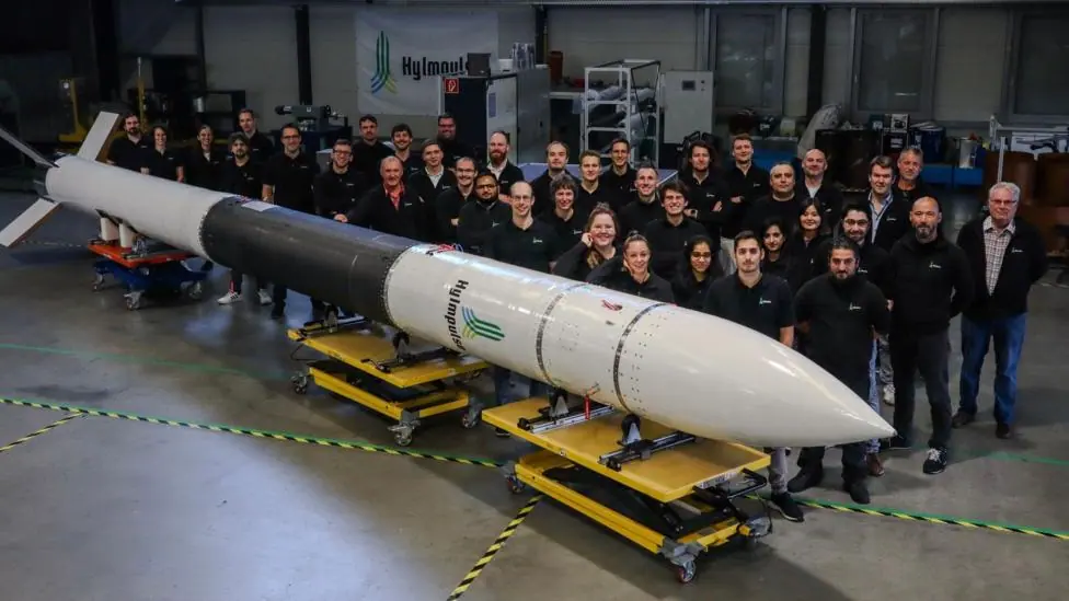 A long and thin white and black rocket lying on its side. A group of about thirty workers are gathered to the right. They're smiling at the camera and standing inside a warehouse.