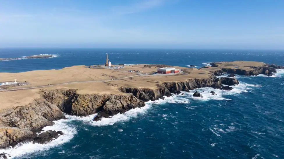 An aerial view of a long flat peninsula surrounded by dark blue sea. The coastline is rocky and the sky is blue. The island is brown and sparsely populated by several industrial buildings and a tall…