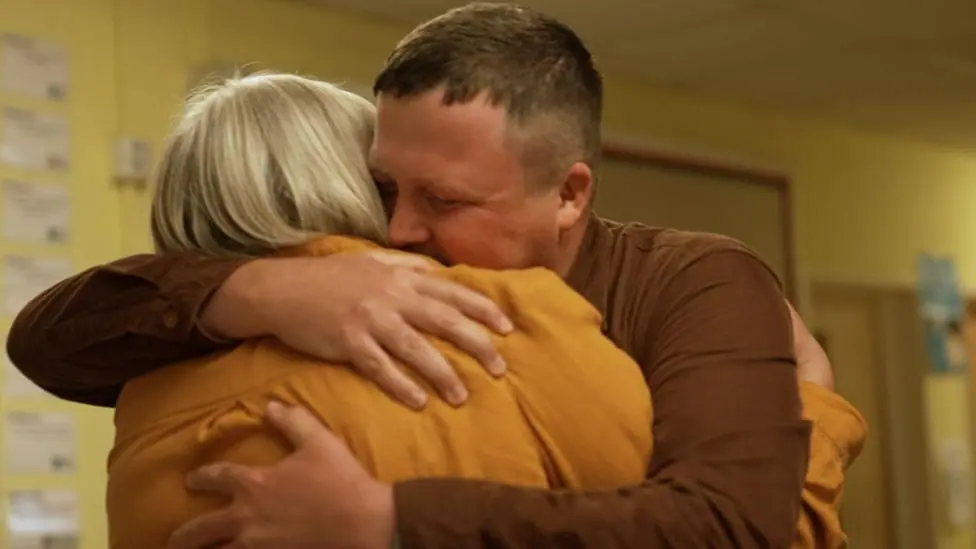 A white man with short brown hair wearing a brown shirt hugs a woman with short blonde hair wearing a dark yellow shirt in a corridor with yellow walls