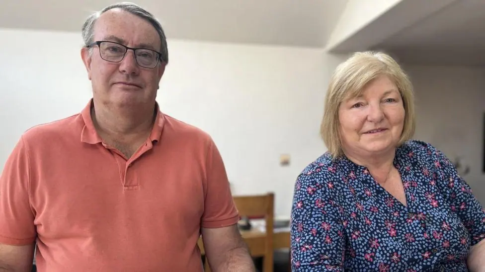 A man in late middle age with short grey hair and wearing a coral-coloured polo shirt and glasses sits at a kitchen island next to a woman of a similar age, with a short blonde bob and wearing a blue…