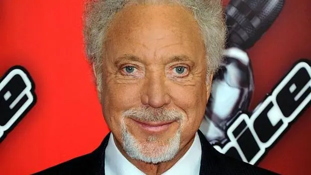 Sir Tom Jones in a blue suit stood in front of a red backdrop with the words The Voice UK on it.