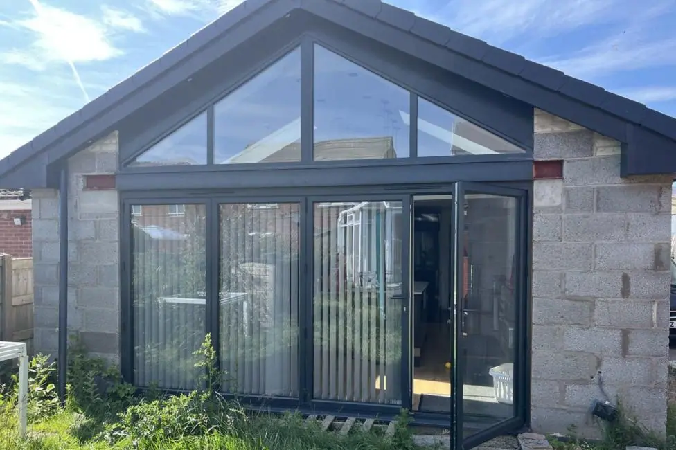 The rear of a bungalow with a pitched cathedral roof and grey bi-fold doors with vertical blinds, taken from outside. The walls are grey breeze block and un-rendered, the grass on the lawn is unkempt…