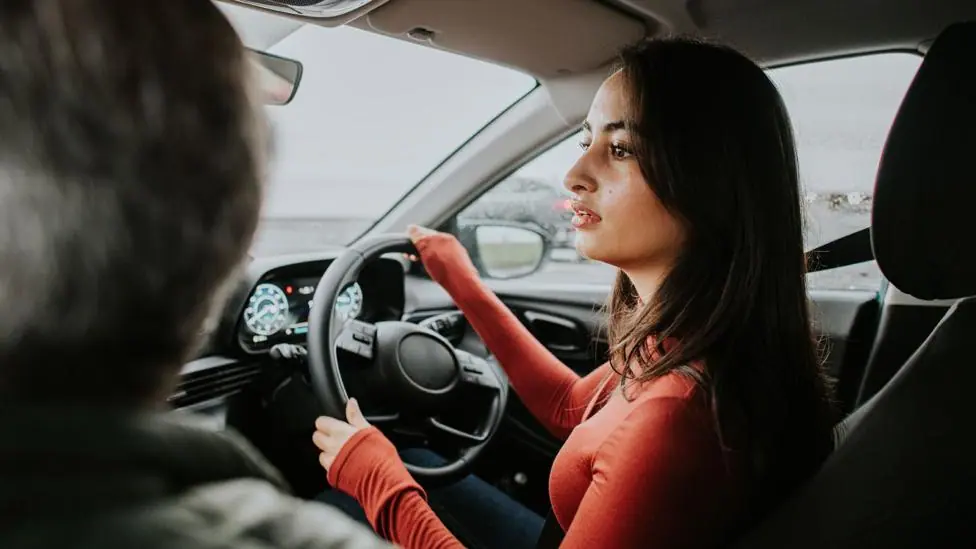 Stock image shows a young person turning a steering wheel as they reverse in a car while an instructor (out of frame) sits in the passenger seat.