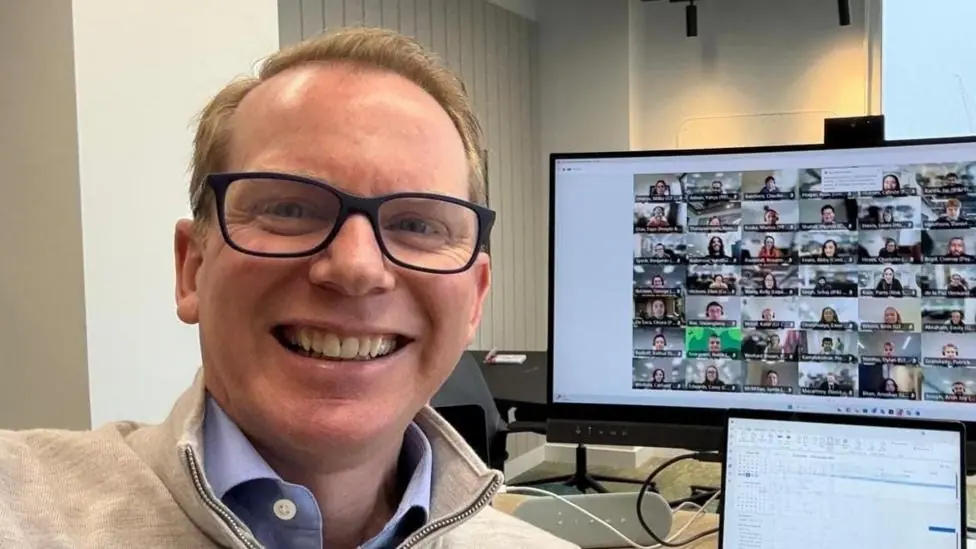 Charlie Nunn, wearing black framed glasses, smiles in a selfie, taken in front of his computer screen where you can see people on a video call
