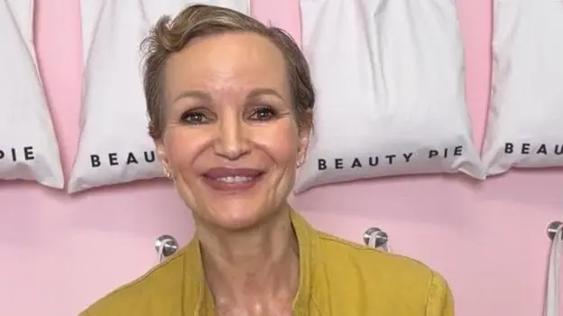 Marcia Kilgore with cropped short brown hair smiling in front of a row of white bags with the Beauty Pie logo on them