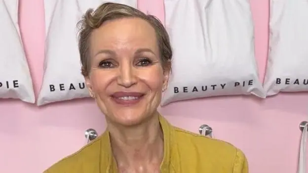 Marcia Kilgore with cropped short brown hair smiling in front of a row of white bag with the Beauty Pie logo on thems at the camera with a blurred background wearing a nude coloured round neck top