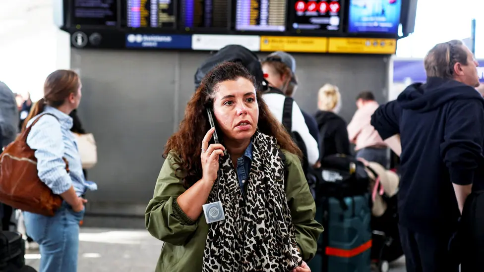A woman wearing a green coat and leopard print scarf talks on the phone inside an airport with flight information on TV screens behind her