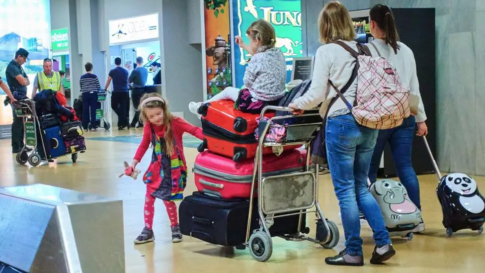 A family with two small children push a luggage trolley pilled with red and pink suitcases through an airport . One of the chidlren is sitting on top of a suitcase and being pushed through the…