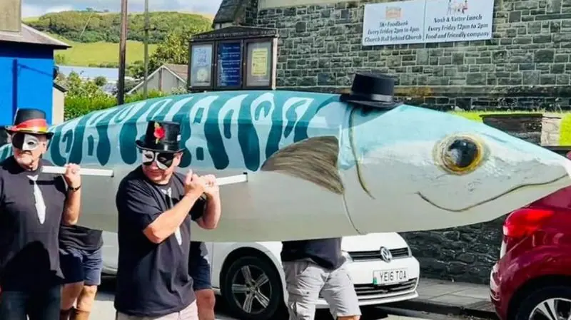 A large blue and white paper mache mackerel. A small black hat is placed on top of the mackerel. It is being carried by six bearers who are wearing black tops with a mackerel printed in the centre…