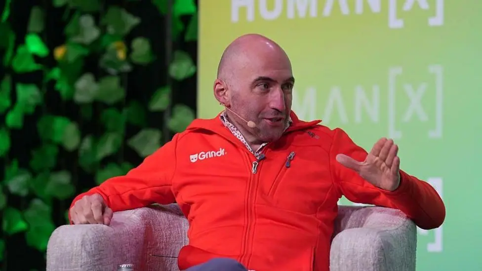 George Arison speaks onstage during the "AI, Identity, and the Future of Connection" panel at the HumanX Conference San Francisco 2026 . He wears a red casual zip up sweater with the Grindr logo in…