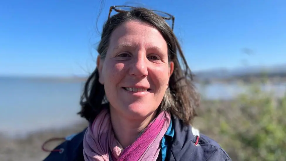 Dr Linda Osti, smiling at the camera with a coastal view behind her. She has sunglasses on her head and is wearing a blue coat with a pink scarf.