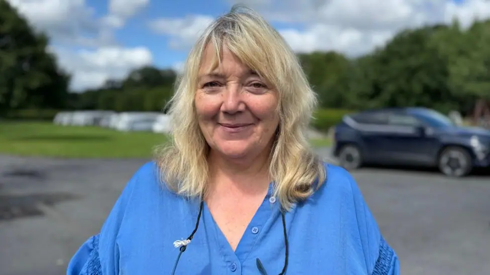 Bethan, who has long blonde hair and is wearing a blue blouse. She is smiling and looking at the camera with a car park and green field with caravans on it behind her.