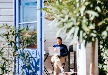 A woman in a blue jumper sits working on a laptop in a blue garden shed.